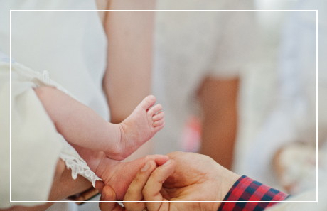 Billede af mennesker klædt i hvid, hvor en voksen hånd holder en babys fod - Dåbs billede